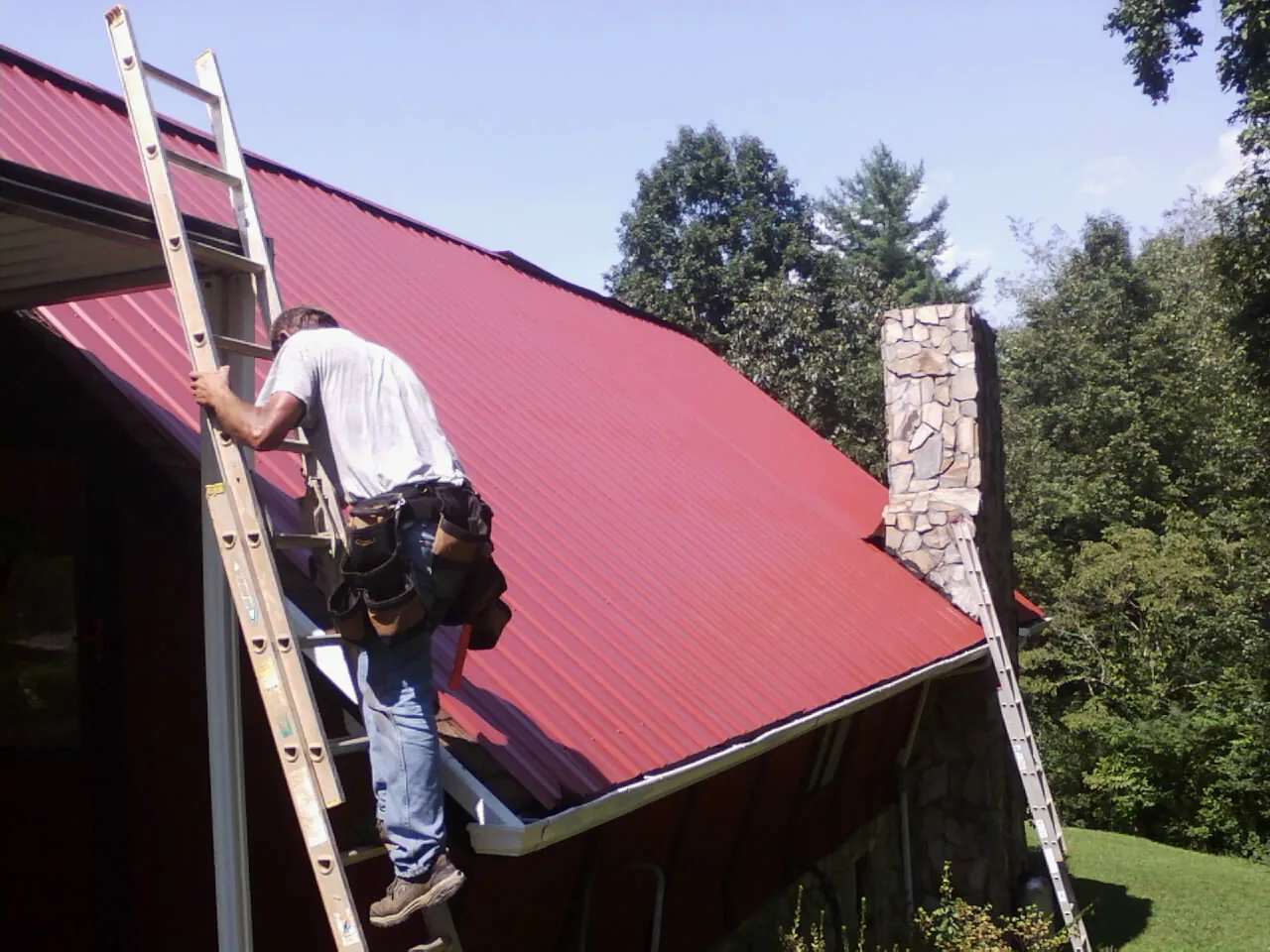 Roofer on a ladder beside a red metal roof and stone chimney — Soffit & Fascia Repair by Roofing Brothers in Stanford